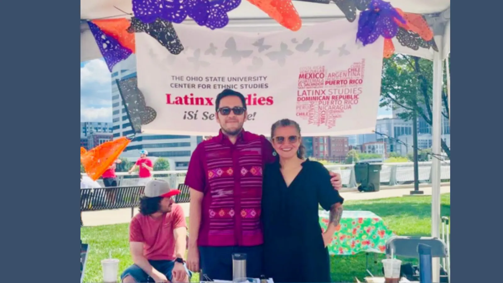A man and a woman stand at a table, surrounded by papel picado paper flags.