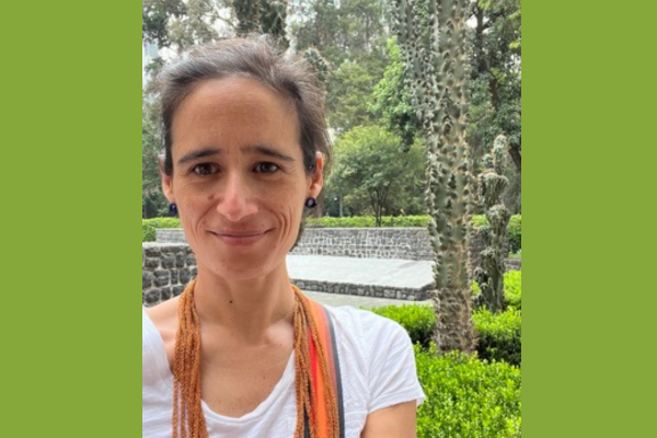 Headshot of Ines Valdez. She has brown hair that is pulled back, is wearing a white vneck tshirt and an orange scarf.