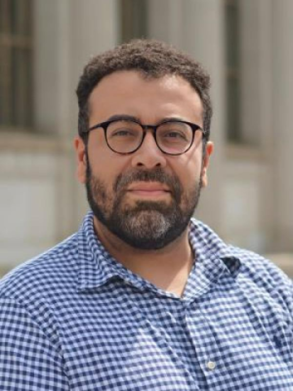 A front portrait of a man with a brown beard, brown hair and round brown glasses. He is wearing a blue checked buttondown shirt. 